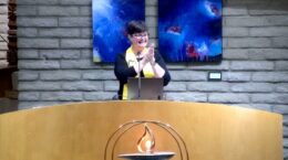 Smiling Rev Christine in the Sanctuary, hands clasped, lit chalice in front of the pulpit